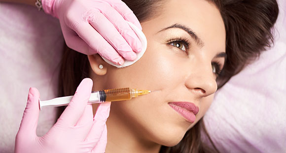 a brunette woman receiving a dermal filler injection to the cheek from a pink-gloves beautician